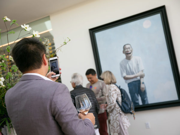 A man takes a photograph of a painting while holding a glass of wine.