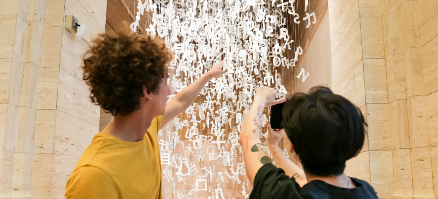 Two people pointing at white ceramic letters hanging from the ceiling.